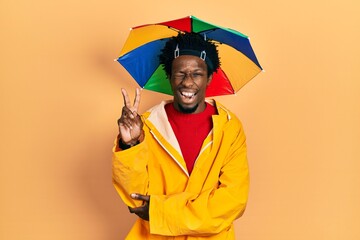 Young african american man wearing yellow raincoat smiling with happy face winking at the camera doing victory sign with fingers. number two.