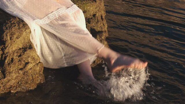 Woman in a white dress dangling water with legs at sunrise. Relaxing and touching sea with feet water from the rock at sunset. 
