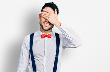 Hispanic man with beard wearing hipster look with bow tie and suspenders smiling and laughing with hand on face covering eyes for surprise. blind concept.
