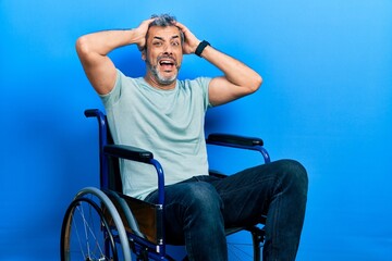 Fototapeta premium Handsome middle age man with grey hair sitting on wheelchair crazy and scared with hands on head, afraid and surprised of shock with open mouth