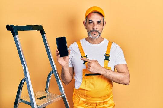 Handsome middle age man with grey hair standing by ladder showing smartphone relaxed with serious expression on face. simple and natural looking at the camera.