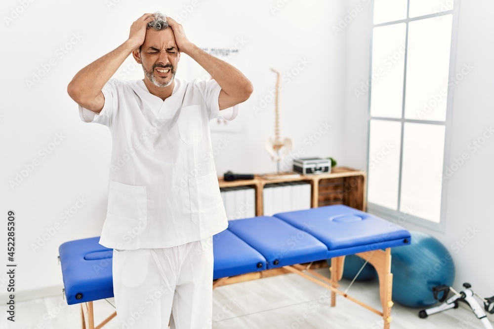 Sticker Middle age hispanic therapist man working at pain recovery clinic suffering from headache desperate and stressed because pain and migraine. hands on head.