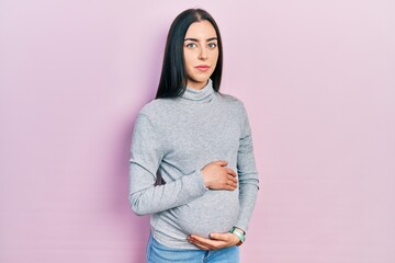 Beautiful woman with blue eyes expecting a baby, touching pregnant belly relaxed with serious expression on face. simple and natural looking at the camera.