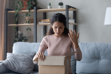 Angry Latin woman sit on sofa unpacking parcel feels dissatisfied while looks inside of cardboard...