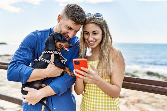 Young caucasian couple using smartphone standing with dog at the beach.