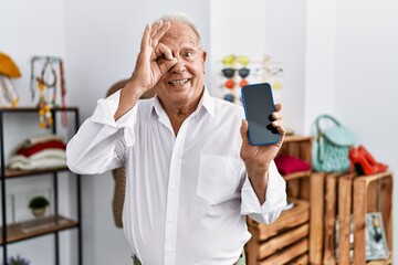 Senior man holding smartphone at retail shop smiling happy doing ok sign with hand on eye looking through fingers