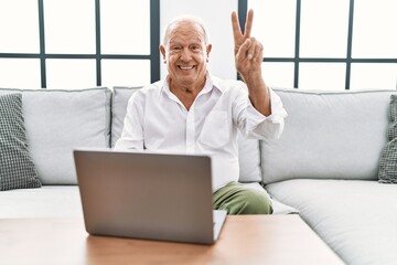 Senior man using laptop at home sitting on the sofa smiling looking to the camera showing fingers...