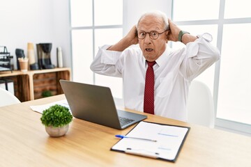 Senior man working at the office using computer laptop crazy and scared with hands on head, afraid and surprised of shock with open mouth
