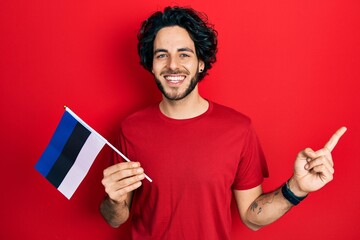 Handsome hispanic man holding estonia flag smiling happy pointing with hand and finger to the side