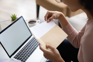 Close up young businesswoman opens envelope with correspondence, takes out letter the latest news seated near laptop with white mock up screen. Paperwork, business notice, invitation to event concept
