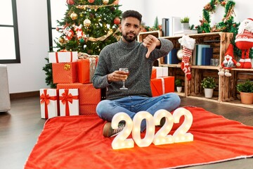 Young hispanic man with beard sitting by christmas tree celebrating 2022 new year with angry face,...