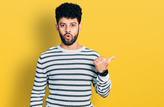 Young arab man with beard wearing casual striped sweater surprised pointing with hand finger to the side, open mouth amazed expression.