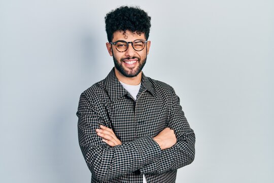 Young arab man with beard wearing glasses with arms crossed gesture winking looking at the camera with sexy expression, cheerful and happy face.
