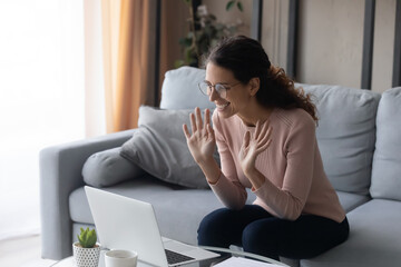 Happy young Latina woman sit on sofa at home smile looks at laptop screen wave hands greeting friend start video call on-line conversation. Modern tech, remote communication, virtual meeting concept