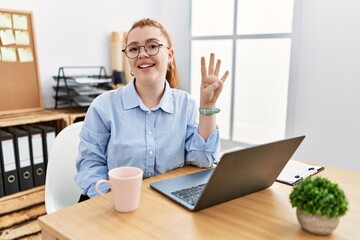 Young redhead woman working at the office using computer laptop showing and pointing up with fingers number four while smiling confident and happy.