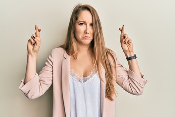 Young blonde woman wearing business jacket and glasses gesturing finger crossed smiling with hope and eyes closed. luck and superstitious concept.