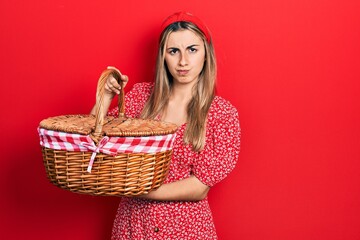 Beautiful hispanic woman holding picnic wicker basket skeptic and nervous, frowning upset because...