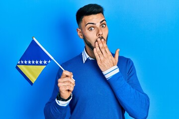 Young hispanic man with beard holding bosnia herzegovina flag covering mouth with hand, shocked and afraid for mistake. surprised expression