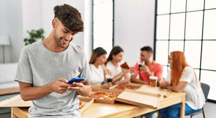 Group of young people eating italian pizza sitting on the table. Man smiling happy using smartphone at home.