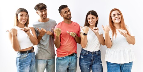 Obraz premium Group of young friends standing together over isolated background pointing to the back behind with hand and thumbs up, smiling confident