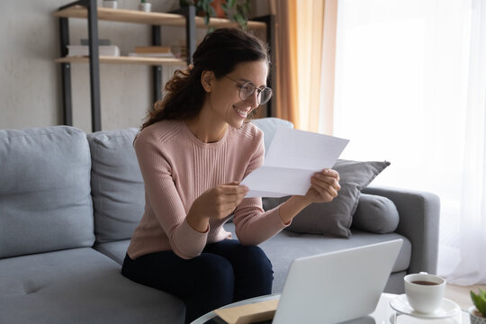 Lovely young woman in glasses holding sheet get acquainted with the latest news read letter feels satisfied pleased sit on sofa at home. Bank notification, last payment, repayment, good news concept