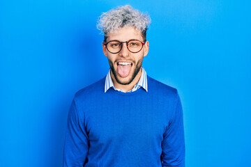 Young hispanic man with modern dyed hair wearing sweater and glasses sticking tongue out happy with funny expression. emotion concept.