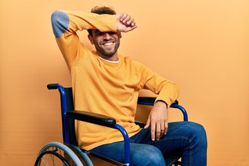 Handsome man with beard sitting on wheelchair covering eyes with arm smiling cheerful and funny. blind concept.