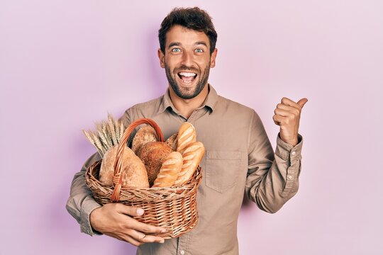 Handsome man with beard holding wicker basket with bread pointing thumb up to the side smiling happy with open mouth