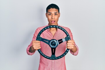 Young african american guy holding steering wheel afraid and shocked with surprise and amazed expression, fear and excited face.