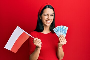 Young hispanic woman holding poland flag and zloty banknotes winking looking at the camera with...