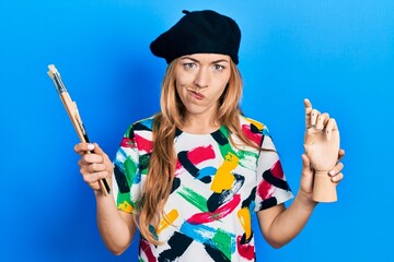 Young caucasian woman artist holding small wooden manikin and paintbrushes skeptic and nervous,...