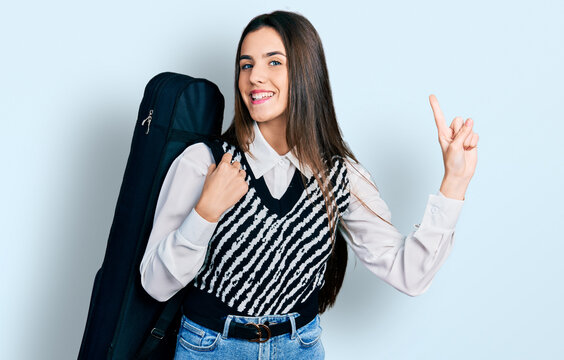 Young brunette teenager wearing guitar case smiling with an idea or question pointing finger with happy face, number one