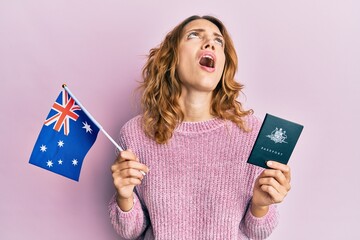 Young caucasian woman holding australian flag and passport angry and mad screaming frustrated and furious, shouting with anger looking up.