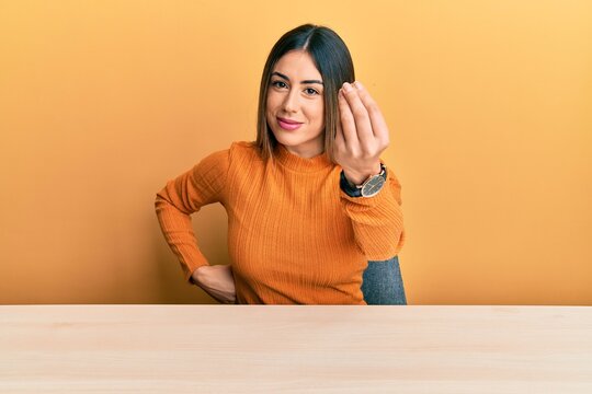 Young hispanic woman wearing casual clothes sitting on the table doing italian gesture with hand and fingers confident expression