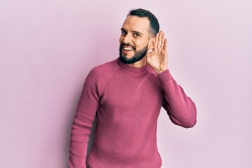 Young man with beard wearing casual winter sweater smiling with hand over ear listening and hearing to rumor or gossip. deafness concept.