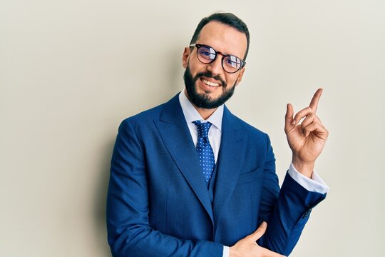 Young man with beard wearing business suit and tie with a big smile on face, pointing with hand and finger to the side looking at the camera.