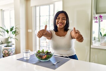 Obraz na płótnie Canvas Young hispanic woman eating healthy salad at home approving doing positive gesture with hand, thumbs up smiling and happy for success. winner gesture.