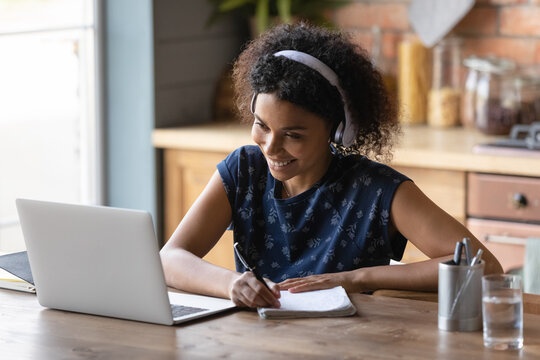 Happy African American millennial student girl wearing earphones studying from home, watching learning webinar, virtual training, online video lesson on laptop, writing notes at kitchen table