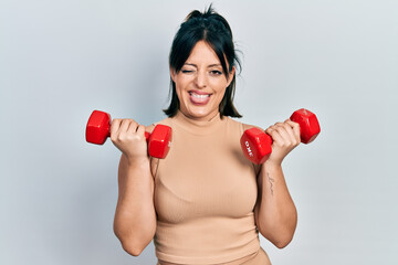 Young hispanic woman wearing sportswear using dumbbells winking looking at the camera with sexy...