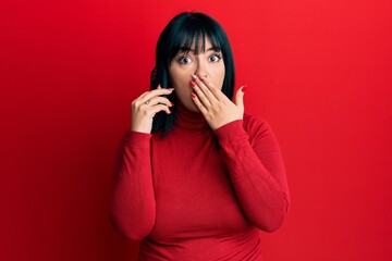 Fototapeta premium Young hispanic woman having conversation talking on the smartphone covering mouth with hand, shocked and afraid for mistake. surprised expression
