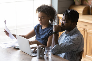Serious African American married couple reviewing monthly bills, analyzing family budget, planning costs. Husband and wife using laptop and reading documents together, filling papers for mortgage
