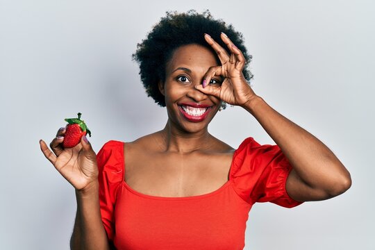 Young African American Woman Holding Strawberry Smiling Happy Doing Ok Sign With Hand On Eye Looking Through Fingers
