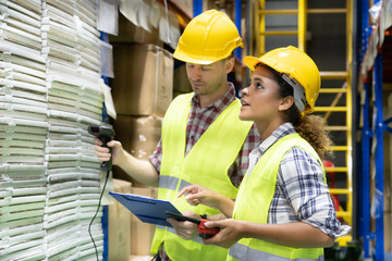 Two warehouse workers doing a stocktaking together in warehouse with African female worker holding a walkie-talkie counting products while Caucasian male holding barcode scanner looking at clipboard.