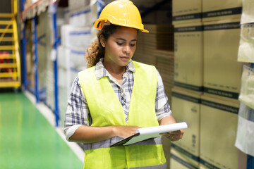 Young pretty African female warehouse worker in a yellow safety vest and helmet standing at a...
