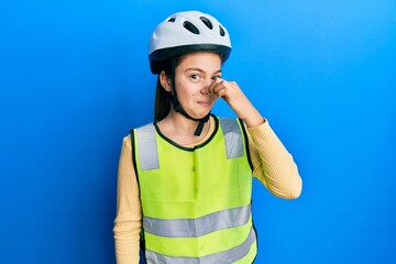 Beautiful brunette little girl wearing bike helmet and reflective vest smelling something stinky and disgusting, intolerable smell, holding breath with fingers on nose. bad smell