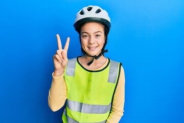 Beautiful brunette little girl wearing bike helmet and reflective vest showing and pointing up with fingers number two while smiling confident and happy.