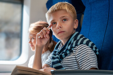 Happy children travels on train returns to home from holidays. Boy prepare to Back to school. Boy thinks about book and looks into distance. Happy girl sits by window in background