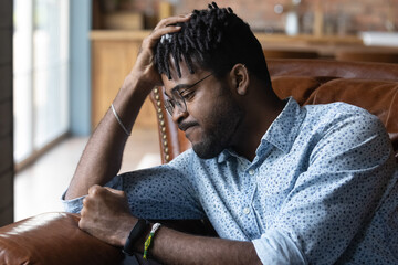Frustrated disappointed Black guy feeling anger, regret about lost opportunities. Young African man suffering from negative mood, depression at home, beating couch with fist