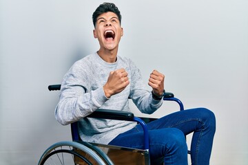 Young hispanic man sitting on wheelchair angry and mad screaming frustrated and furious, shouting...