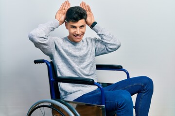 Young hispanic man sitting on wheelchair doing bunny ears gesture with hands palms looking cynical...